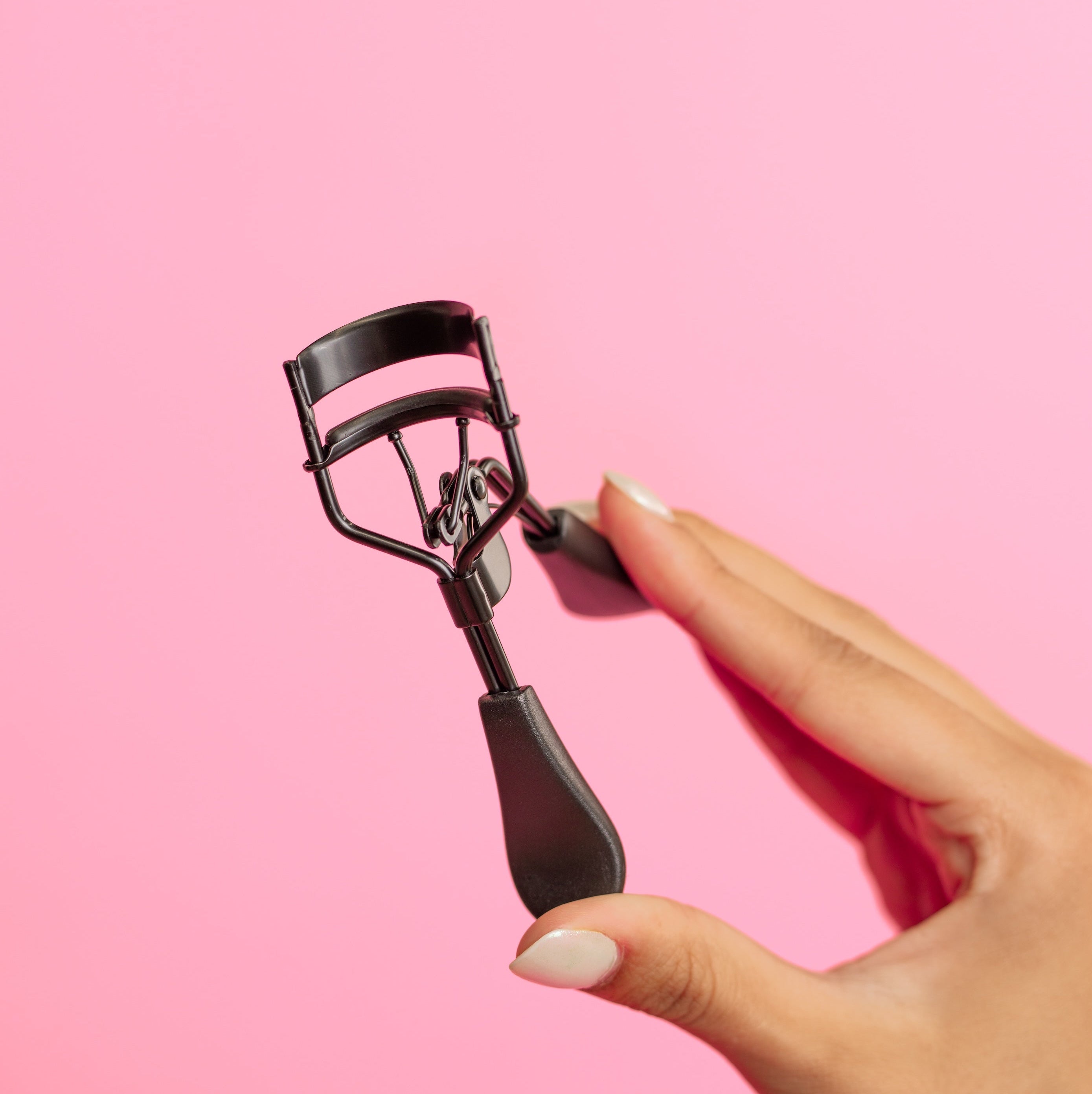 Eyelash curler held by a hand against a pink background